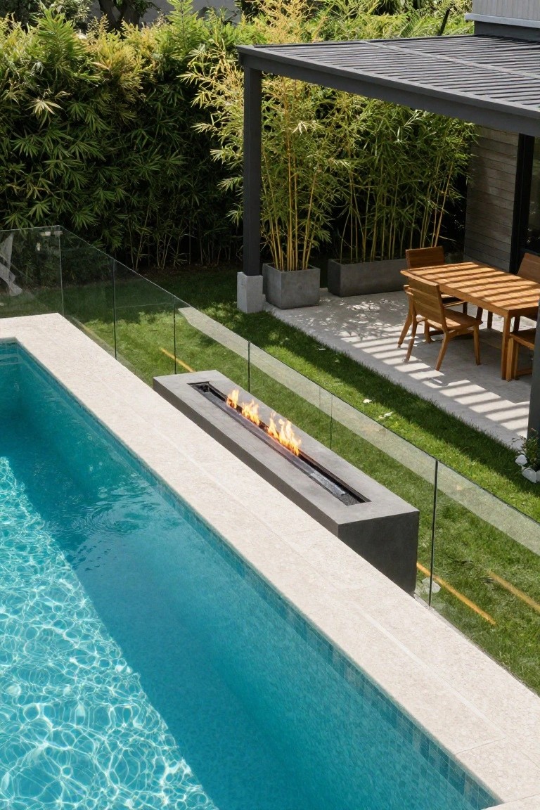 Rectangular swimming pool with a linear gas fire feature along one edge on a concrete deck, blue water, glass pool fencing, bamboo plants, and nearby outdoor dining table with wooden chairs under a covered patio.