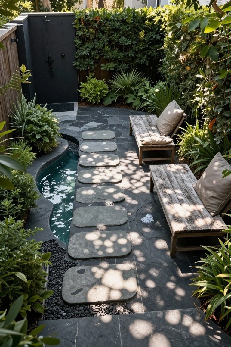Narrow curving inground pool in a lush outdoor courtyard with large flat gray stepping stones across the water, wooden lounge chairs with cushions nearby, tropical plants, black pebble edging, and a black outdoor shower enclosure.