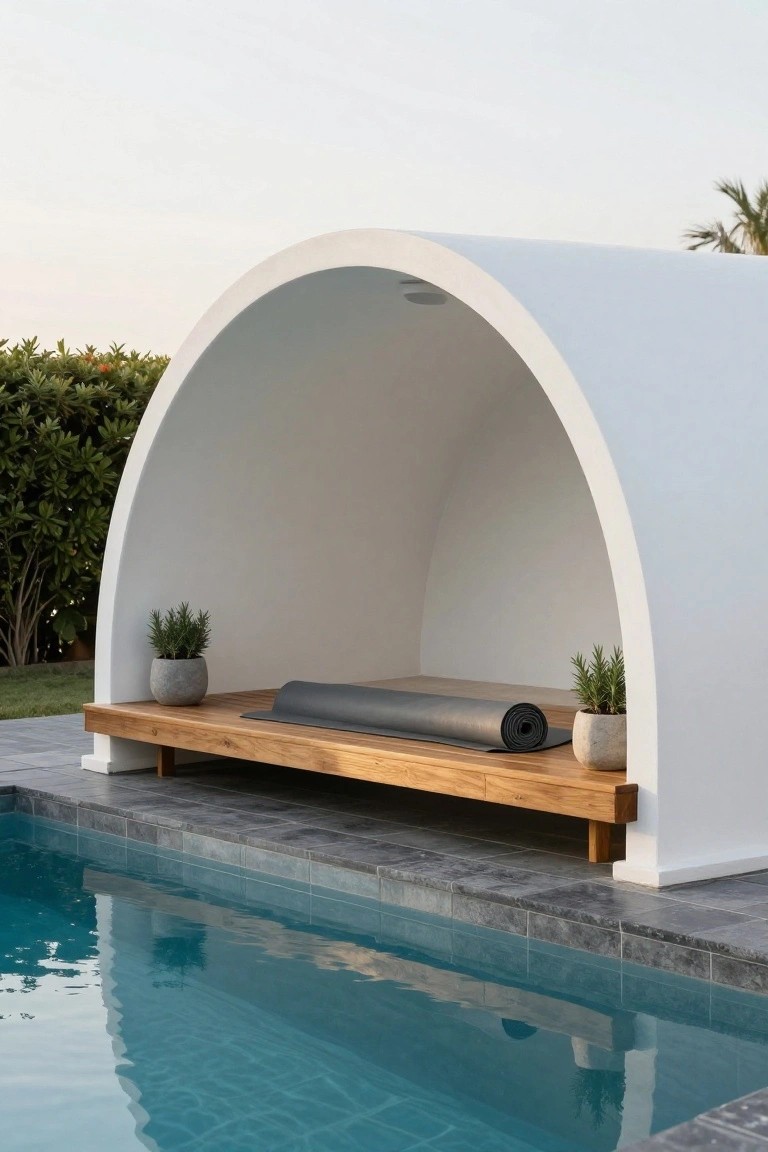 White arched cabana with wooden platform, rolled gray yoga mat, and potted rosemary plants on each side, positioned next to a turquoise pool with dark gray tile edging and surrounded by green shrubs and palms.