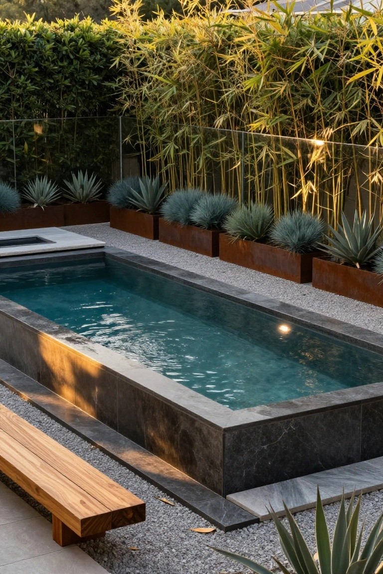 Narrow rectangular pool with dark stone edges and turquoise water, surrounded by pebble gravel, corten steel planters containing agave and succulent plants, bamboo screening in the background, glass fencing, and a long wooden bench alongside.