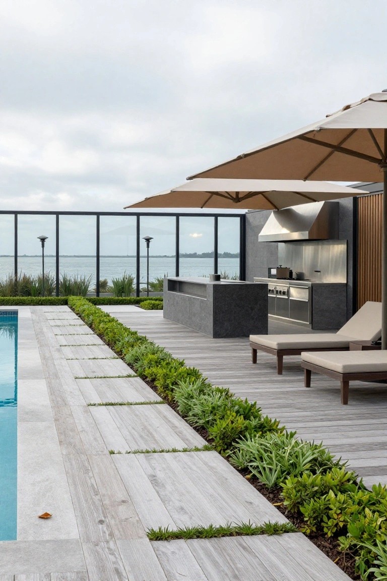 Pool deck with light stone coping, linear pale wood plank pathway along the edge bordered by green plants, two beige loungers, outdoor kitchen with stainless steel hood, glass railing, and water view in background.