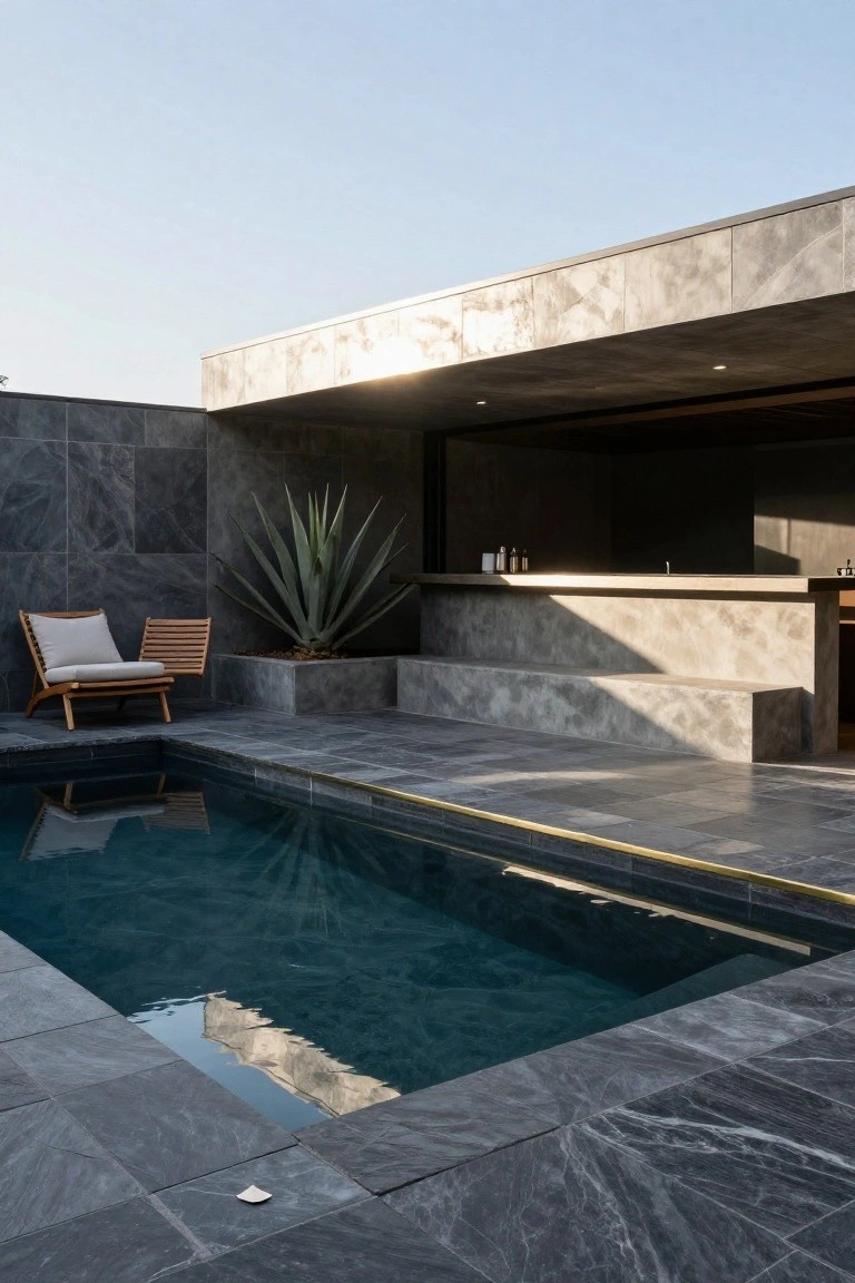 Small rectangular pool with dark veined stone tile surround, wooden lounge chair, potted agave plant, concrete bar counter, and overhanging modern architecture against tiled walls.