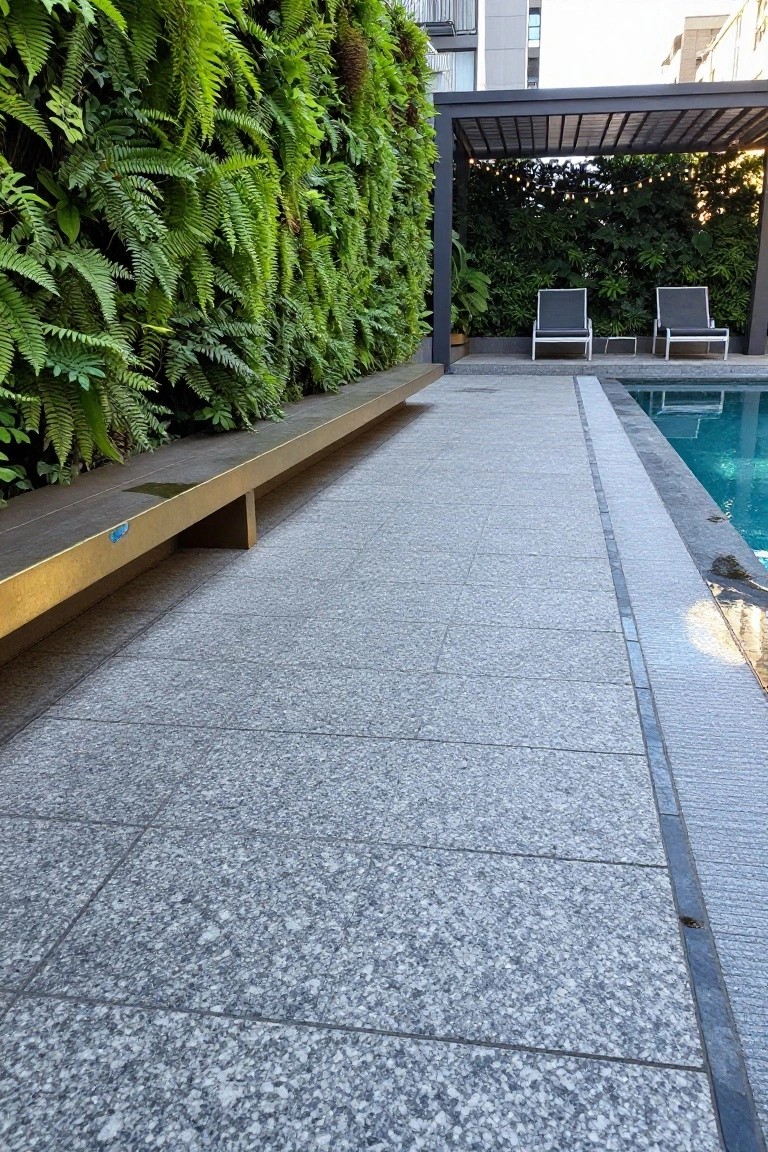 Pool deck paved in light gray granite tiles with a long gold bench, lush green fern wall, two gray lounge chairs under a pergola, and turquoise pool water.