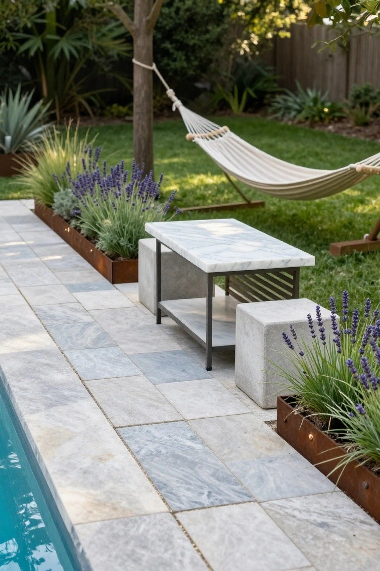 Pool deck paved with light gray marble tiles extending to the turquoise pool edge, with a white hammock, marble-topped metal table, concrete stools, and lavender plants in rusted metal planters on a paved path beside green lawn.