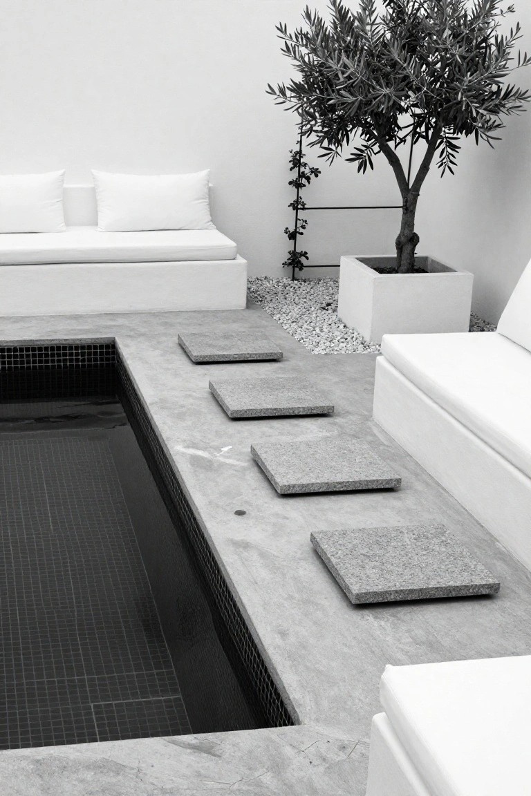 Black mosaic-tiled pool with four square gray granite slabs stepping down from a concrete deck edge, white modular seating nearby, olive tree in white square pot, black and white photo.