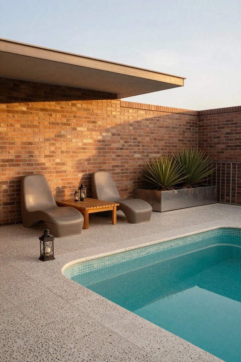 Outdoor pool deck of speckled exposed aggregate concrete featuring two curvy gray lounge chairs around a wooden table, metal lanterns, potted fan palms against a brick wall, and a turquoise tiled pool.