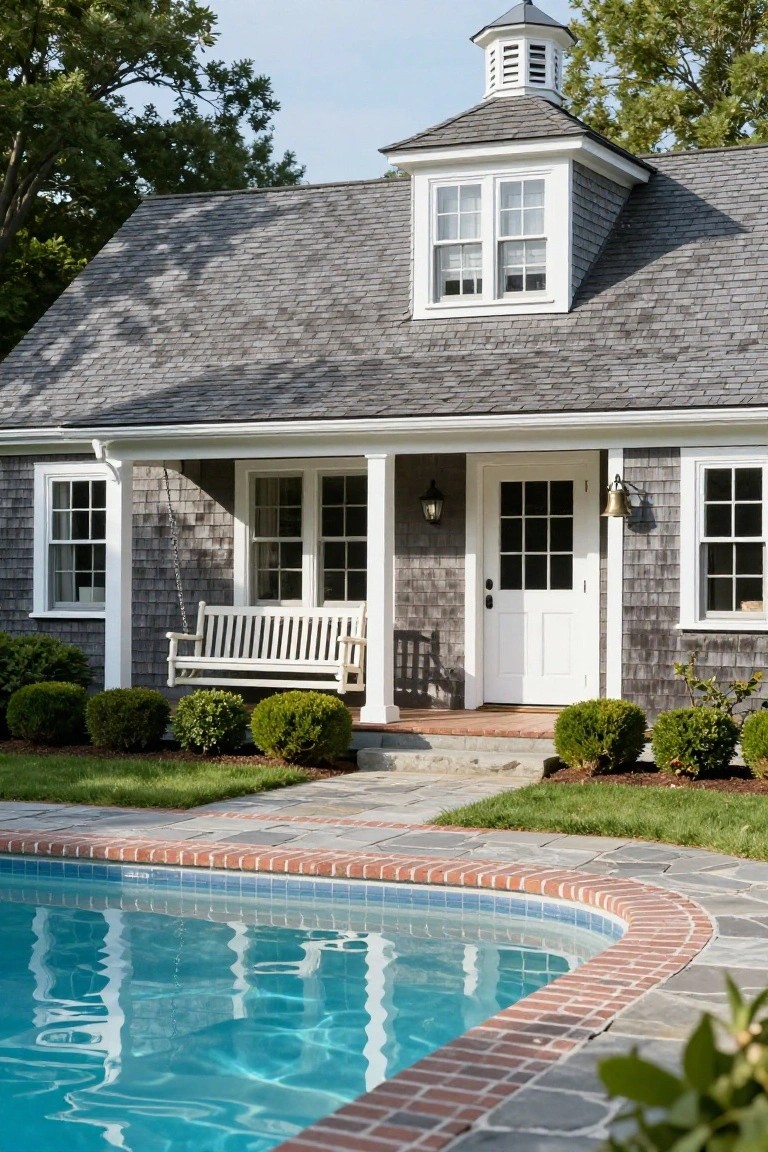 Gray shingle-clad pool house with white-trimmed covered porch, porch swing, lanterns, and boxwoods in front of a curved in-ground pool with brick edging.