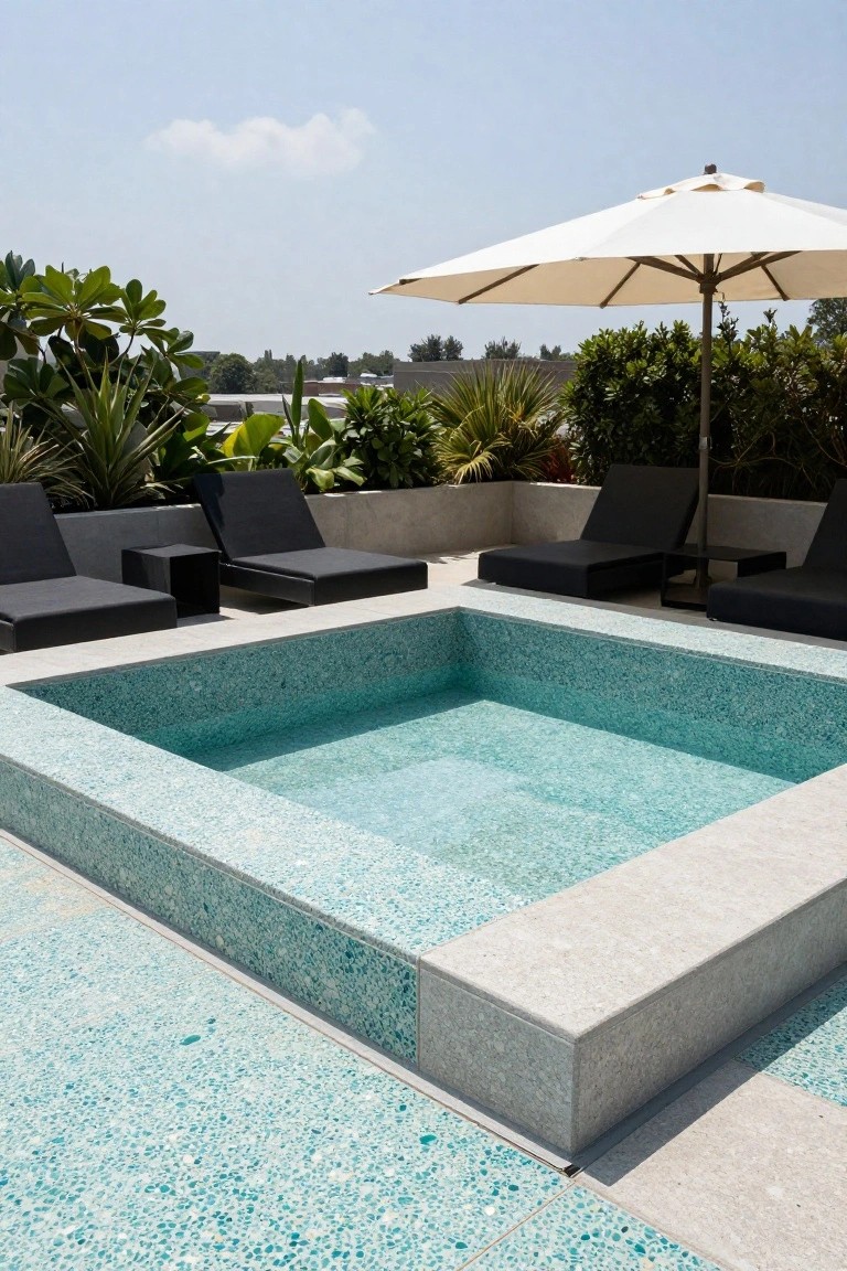 Rooftop pool with turquoise glass mosaic tiled deck and surround, black chaise lounges, side tables, white umbrella, and potted tropical plants against a city skyline backdrop.