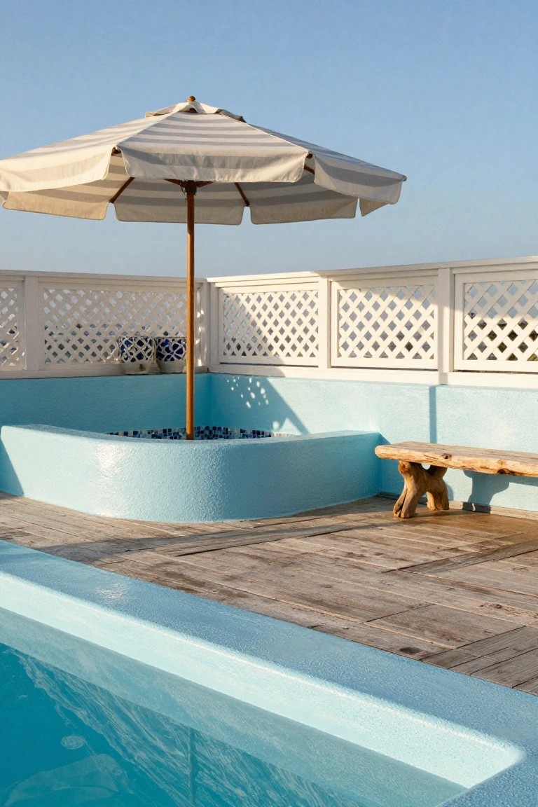 Rooftop pool deck made of weathered wooden planks adjacent to a curved turquoise pool with blue water, featuring a white beach umbrella, blue-tiled fire pit area, wooden bench, and white lattice fencing under a clear sky.