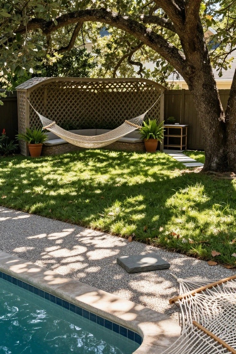 Wooden pergola with beige cushioned hammock beside a turquoise pool, grass lawn, potted plants, and gravel path in a backyard shaded by oak tree.