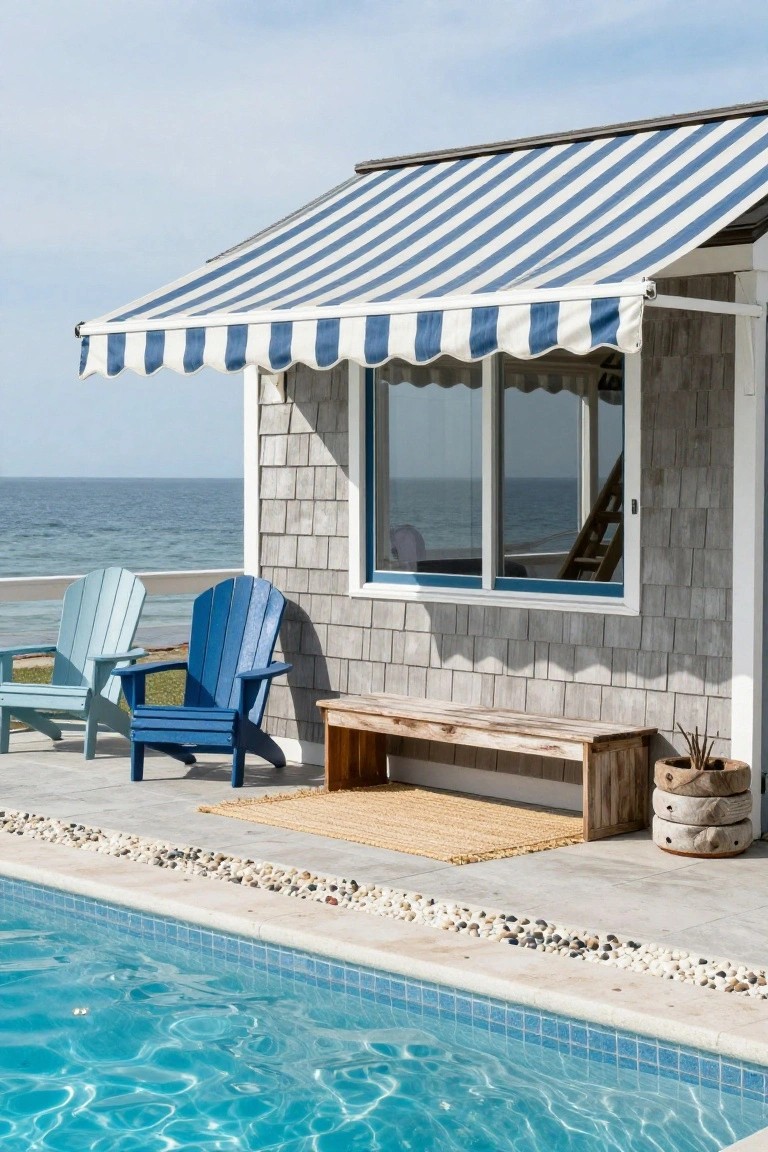 Poolside deck with blue and white striped awning extending from a shingled structure with windows overlooking the ocean, flanked by light blue and dark blue Adirondack chairs, wooden bench, seagrass rug, potted plants, and pebble border next to the pool.