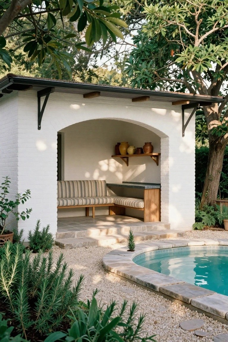 White brick pool house pavilion featuring an arched open entryway with built-in bench seating, wood bar counter, shelves with pottery, adjacent curved swimming pool, gravel ground cover, and surrounding plants and trees.