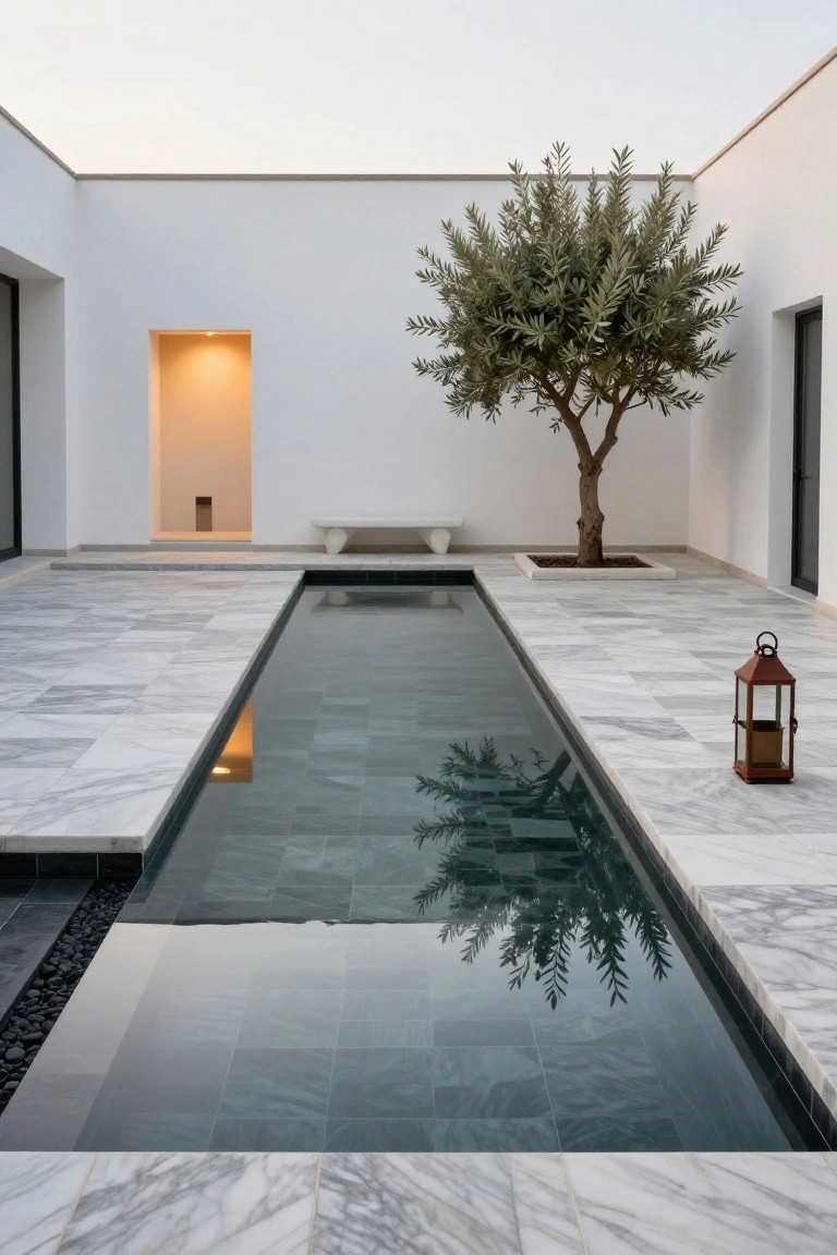Narrow rectangular pool with dark slate geometric tiles and reflections of an olive tree, surrounded by white marble flooring in a minimalist white-walled courtyard with a stone bench and lantern.