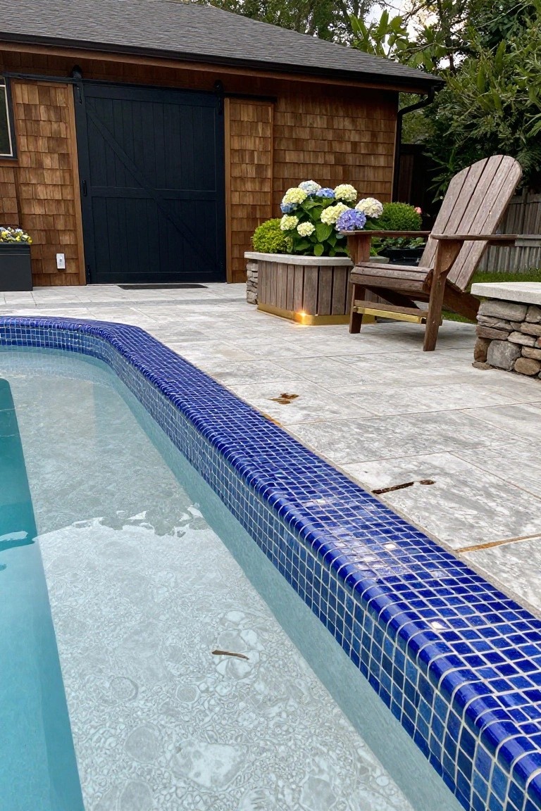 Pool edge tiled with small blue mosaic tiles along the curved border, adjacent to gray stone pavers, a wooden Adirondack chair next to planters with hydrangeas, and a shingle-clad shed nearby.
