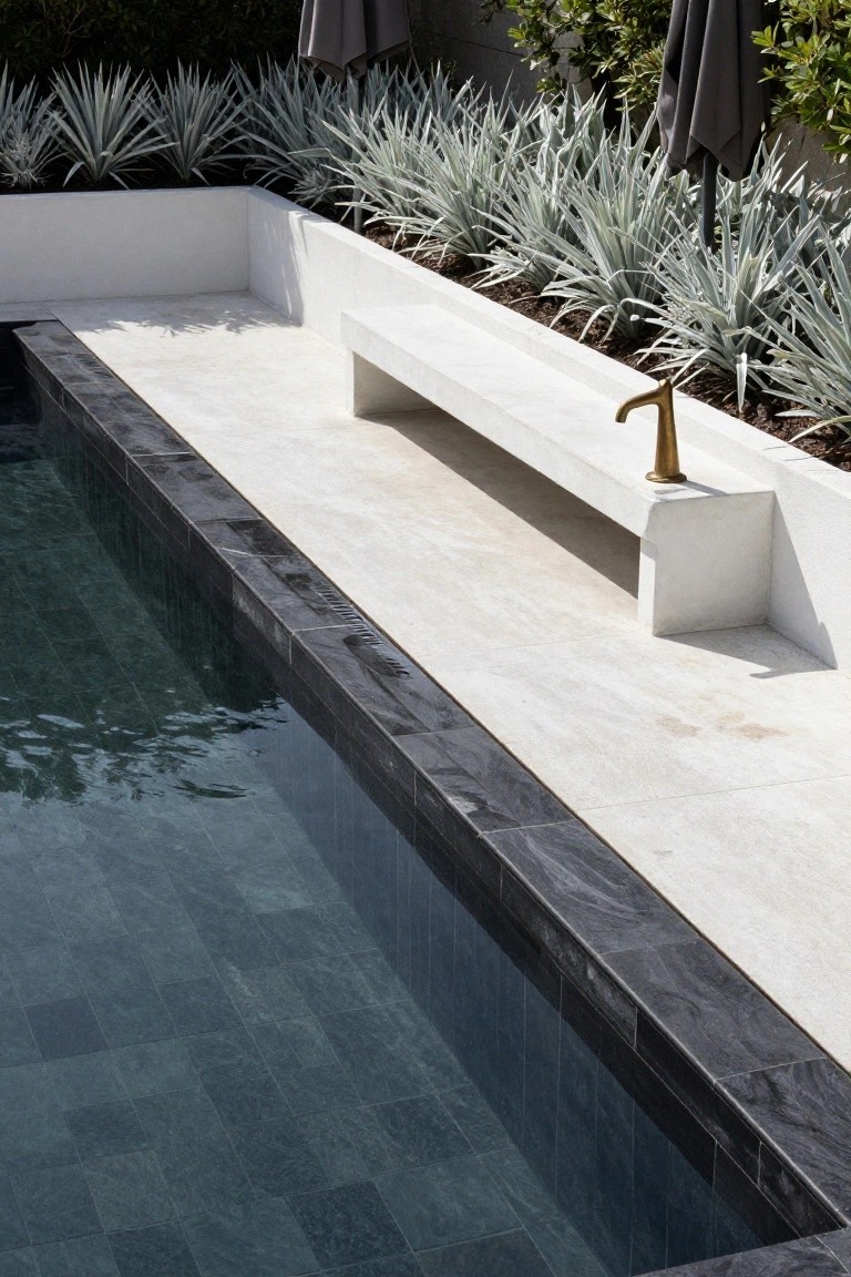 Rectangular pool with dark gray slate tiles, black edge coping, white stone deck and bench, flanked by white agave plants and gray umbrellas against a greenery backdrop.