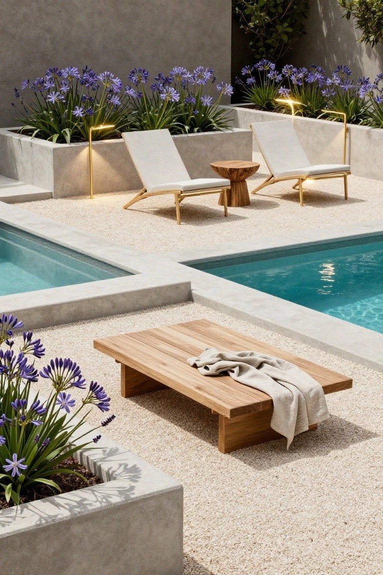 Pool area with concrete planters of purple agapanthus flowers illuminated by tall slim gold lamps, white sling loungers, wooden side table and low coffee table, turquoise pool water, and light gravel ground surrounded by concrete walls.