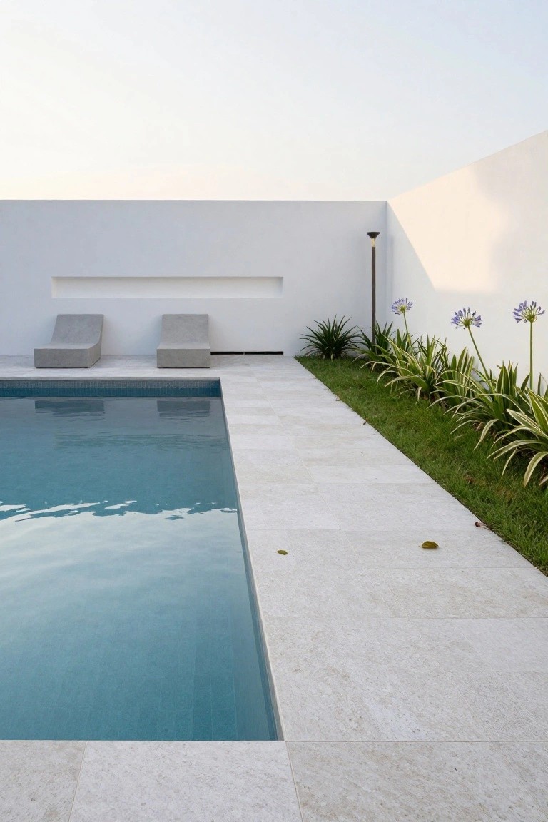 Rectangular turquoise pool with white tiled deck edge, flanked by white walls, two angled gray lounge chairs, green plants, and purple agapanthus flowers along the side.