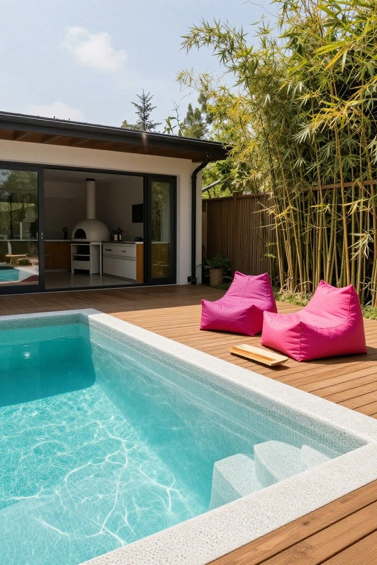24 Relaxing Outdoor Pool Area Ideas For Unwinding After Long Days Wooden deck beside a turquoise infinity-edge pool with white pebble coping and steps, two large pink bean bag chairs with a wooden tray between them, outdoor kitchen with pizza oven visible through glass doors, bamboo privacy screen.