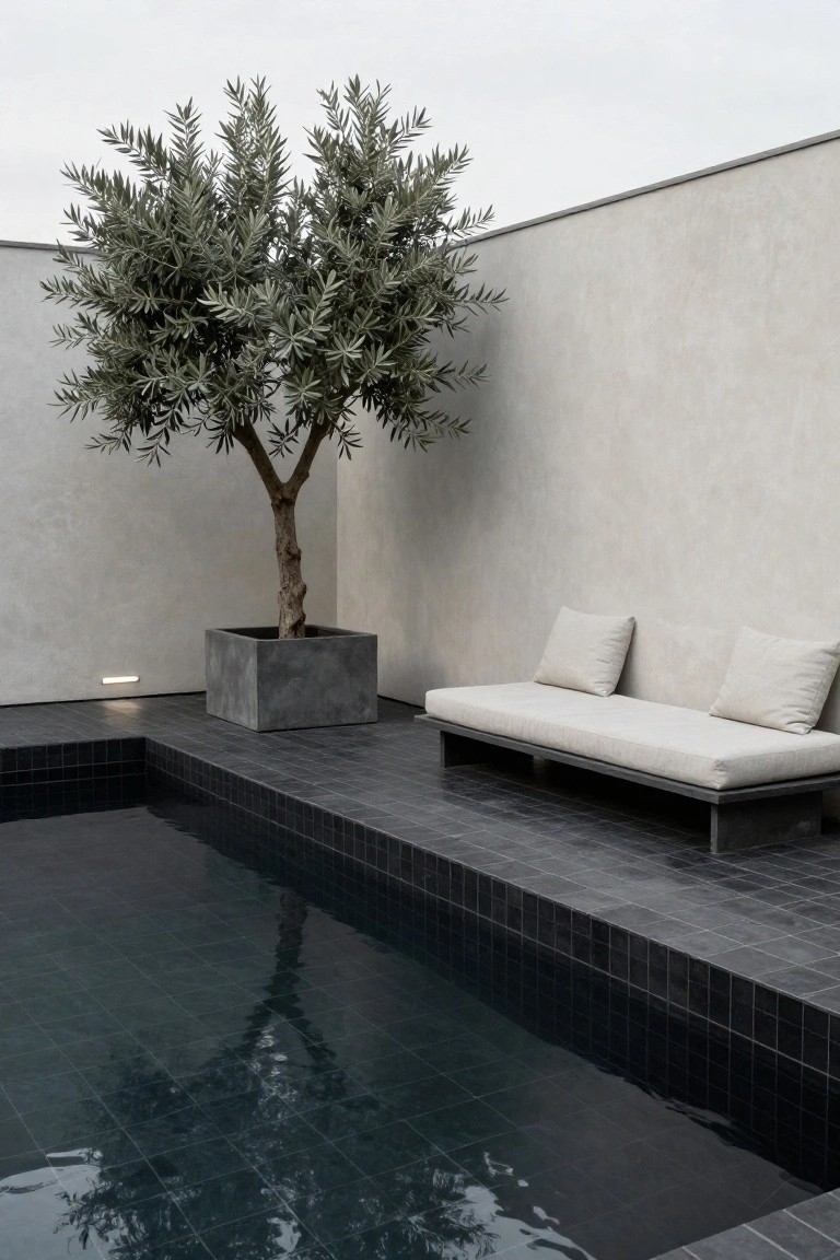 Modern inground pool corner with black mosaic tiles and calm water, low gray metal daybed with beige cushions nearby, large potted olive tree in gray concrete planter, light gray walls, and subtle ground lighting.