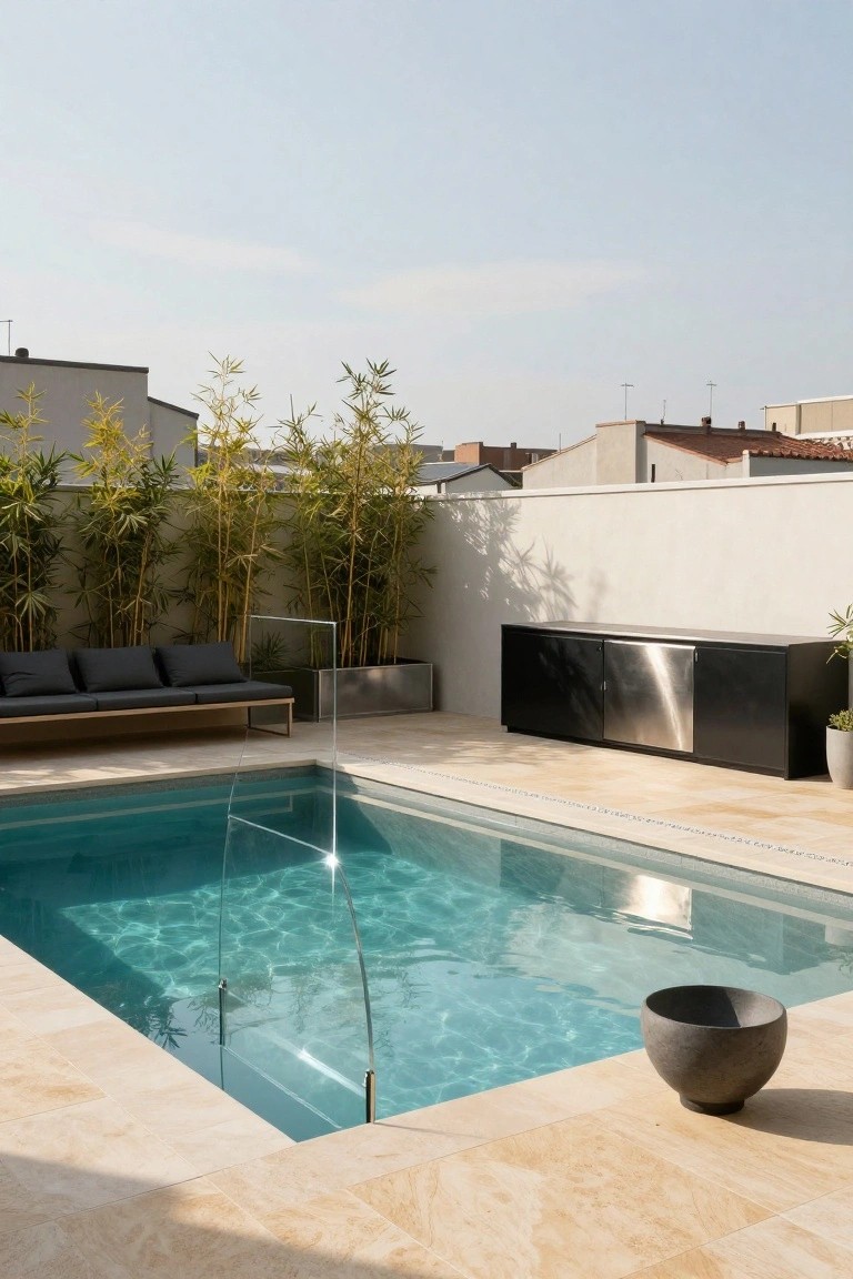 Small rectangular turquoise semi-inground pool with glass fencing and light beige paver surround, black lounge sofa, stainless steel cabinet unit, potted plants, and bamboo screening against a white wall on a terrace.