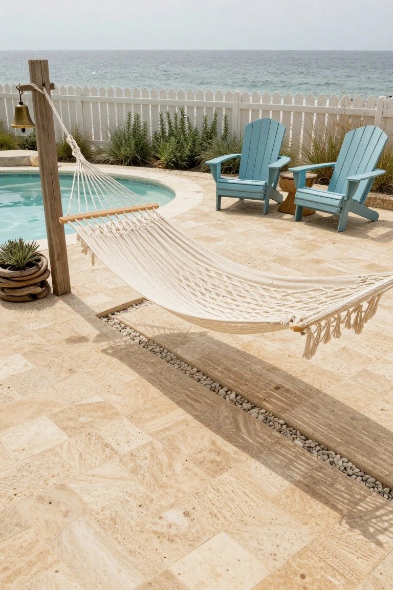 Curved semi-inground pool surrounded by light beige rectangular paver tiles on a patio, with a white rope hammock hanging from wooden posts, two turquoise Adirondack chairs, potted succulent and agave plants, wooden bell post, white picket fence, and ocean in the background.