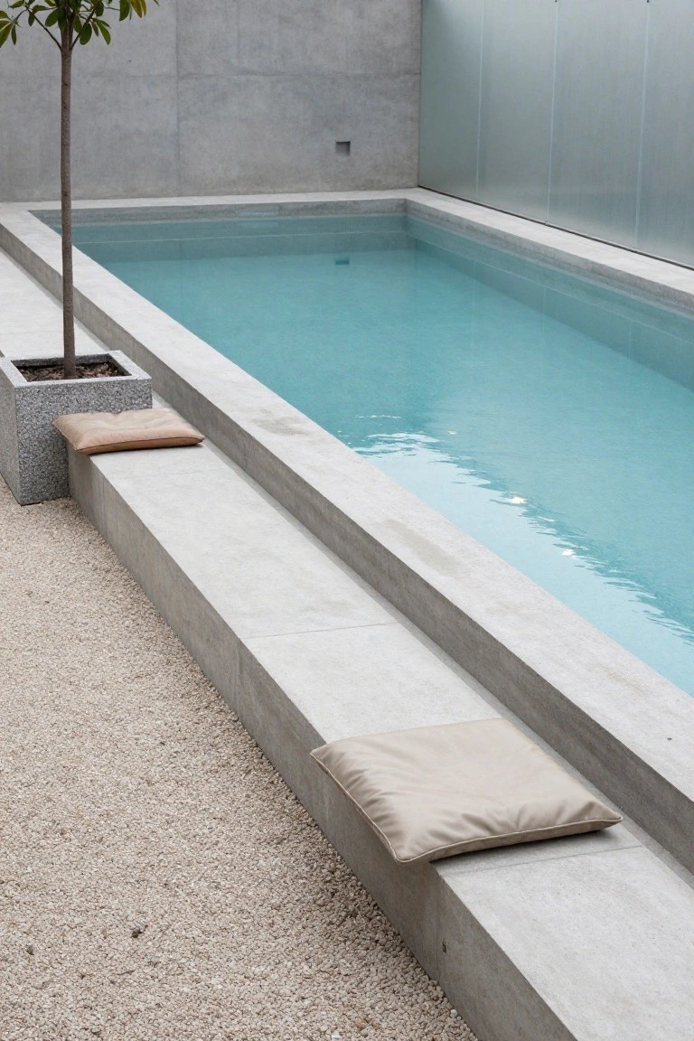 Narrow semi-inground pool with turquoise water next to wide gray concrete steps topped with beige cushions, a potted tree in a stone planter, gravel ground cover, and surrounding concrete walls with glass panels.