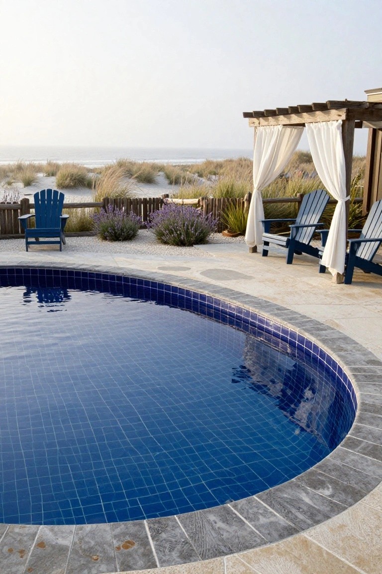 25 Dramatic Dark Blue Pool Tile Ideas With Visual Depth Round outdoor pool with dark blue mosaic tiles and gray stone edging on a light deck, flanked by blue Adirondack chairs under a wooden pergola with white curtains, beach dunes in the background.