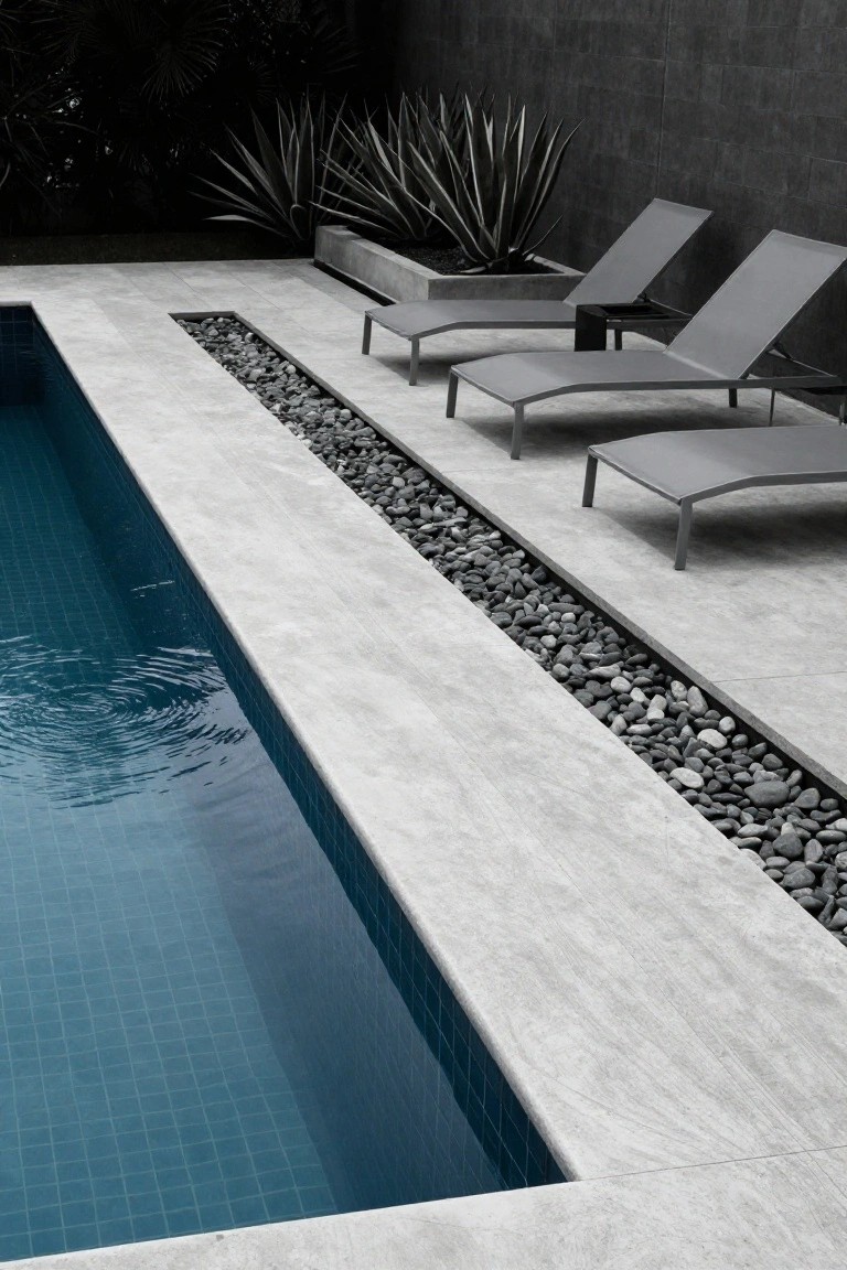 25 Dramatic Dark Blue Pool Tile Ideas With Visual Depth A rectangular pool with dark blue mosaic tiles viewed from the side, featuring a wide concrete deck edged by a linear trench filled with dark pebbles, several grey mesh loungers nearby, agave plants, and a dark textured wall in the background.