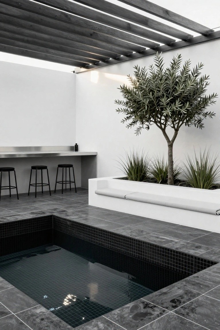 Modern outdoor pool patio with small black mosaic-tiled inground pool, stainless steel bar counter, three black stools, olive tree in planter, white bench, and wooden beam pergola overhead.