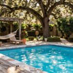 Wooden deck beside turquoise pool with two cushioned lounge chairs, white hammock under pergola hung from large oak tree, potted plants, and stone poolside path.