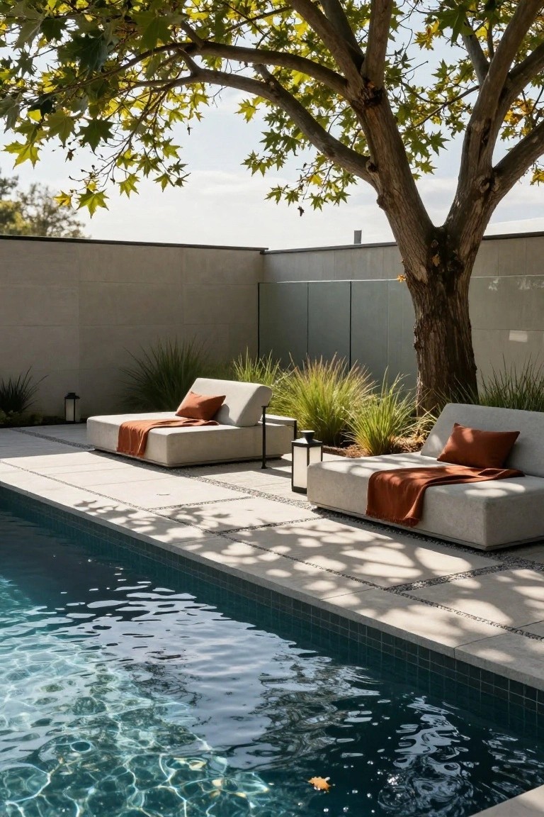 Two white low-profile daybeds with orange cushions and throws positioned under a large tree canopy next to a turquoise-tiled pool, with grasses, lanterns, and a concrete wall nearby.