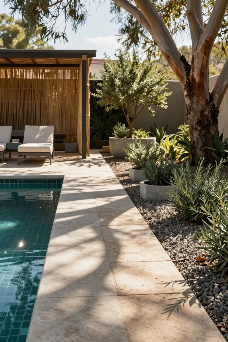 Turquoise-tiled pool edged in light stone pavers, with gravel planting strip holding low silver-leaf shrubs in round concrete pots, tall eucalyptus tree overhanging from above, and two white loungers under bamboo-screened pergola structure.