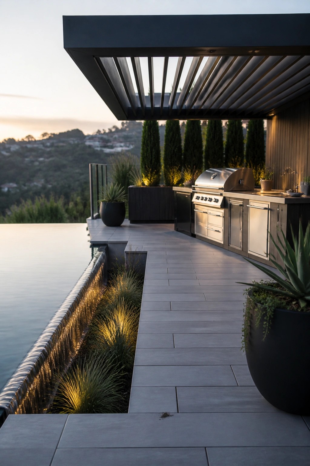 An outdoor poolside patio with a modern kitchen, paved deck, tall potted plants, and a row of narrow evergreen trees in the background.