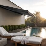 Modern house exterior with a large dark retractable awning extended over a wooden deck beside a long swimming pool, two lounge chairs with white cushions, and a concrete fire pit in the foreground.