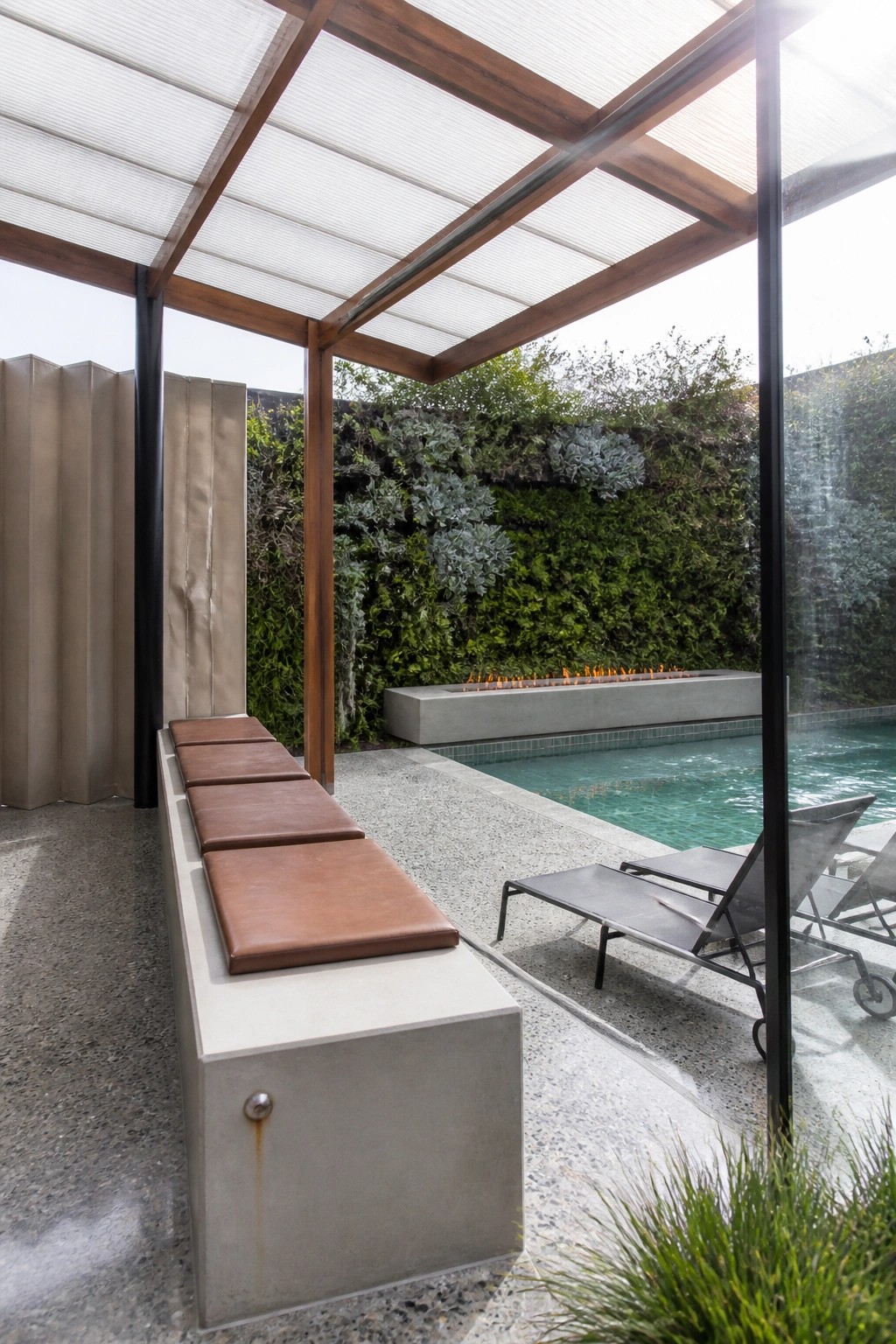 An outdoor pool area with a wooden-framed pavilion featuring a translucent roof, a long concrete bench with cushions, metal lounge chairs, and a living green wall beside the water.