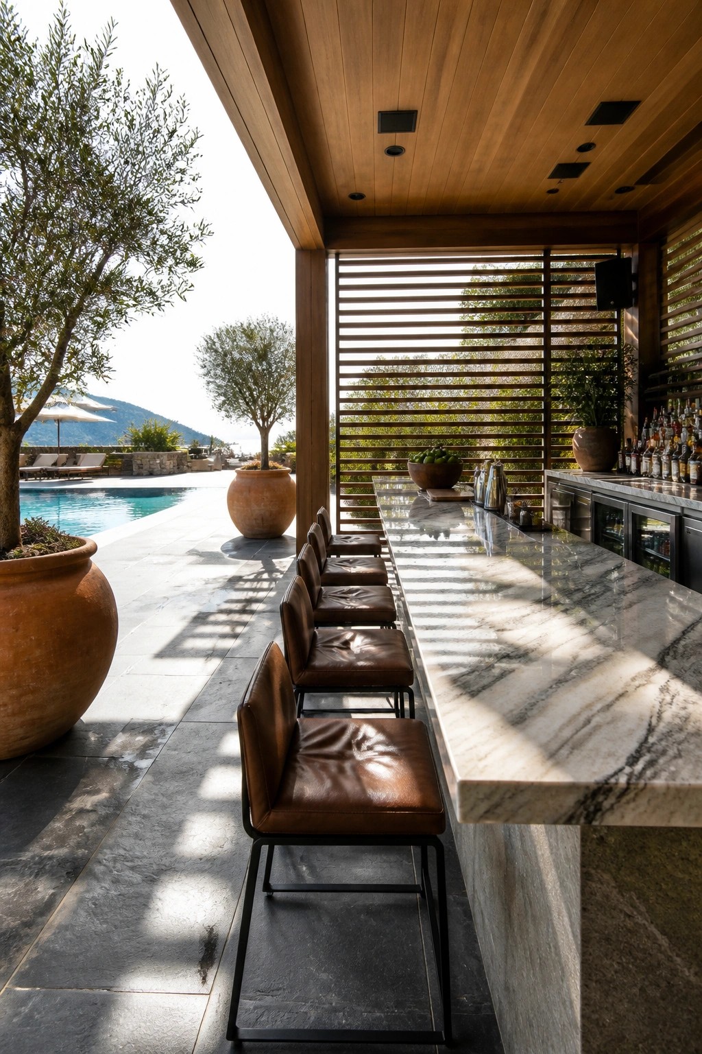 A long marble bar counter with brown leather stools lines an outdoor covered space next to a swimming pool, with trees and hills visible in the background.