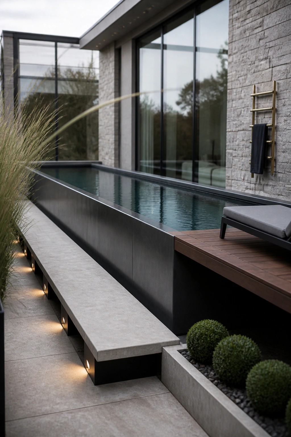 A long rectangular modern pool with a concrete bench featuring integrated lighting running parallel to it, next to a wooden deck and stone house wall.