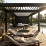 Modern dark pergola over a wooden deck with lounge chairs, fire pit, and side tables next to a pool with glass railings.