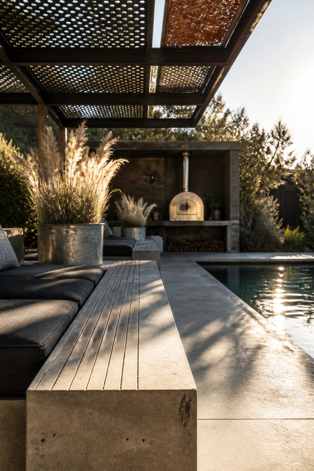 An outdoor poolside patio under a metal pergola with perforated and solid roof panels, featuring concrete benches, potted grasses, a built-in pizza oven, and a swimming pool visible on the right.