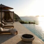 An infinity pool with wooden lounge chairs on a tiled terrace overlooking the ocean, with rolled towels in a stone niche and a fire pit area nearby.
