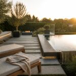 Modern pool with a long cascading water feature along the edge, stone pavers, two lounge chairs in the foreground, and a fire pit area nearby at sunset.