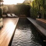A narrow rectangular backyard pool with dark water sits between a wooden bench on the left and a wooden deck on the right, with a fire pit at the far end, lanterns in the foreground, and ivy-covered walls.