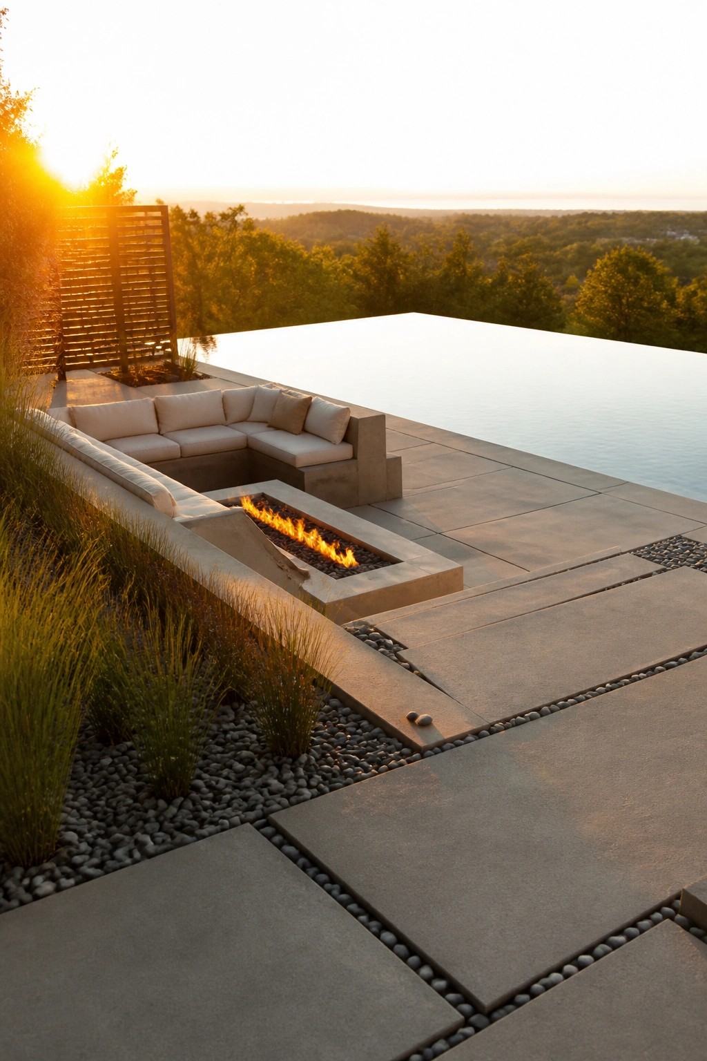 An outdoor pool deck features a rectangular concrete fire pit with built-in cushioned seating on one side and a large infinity pool on the other, surrounded by gravel beds with grasses.