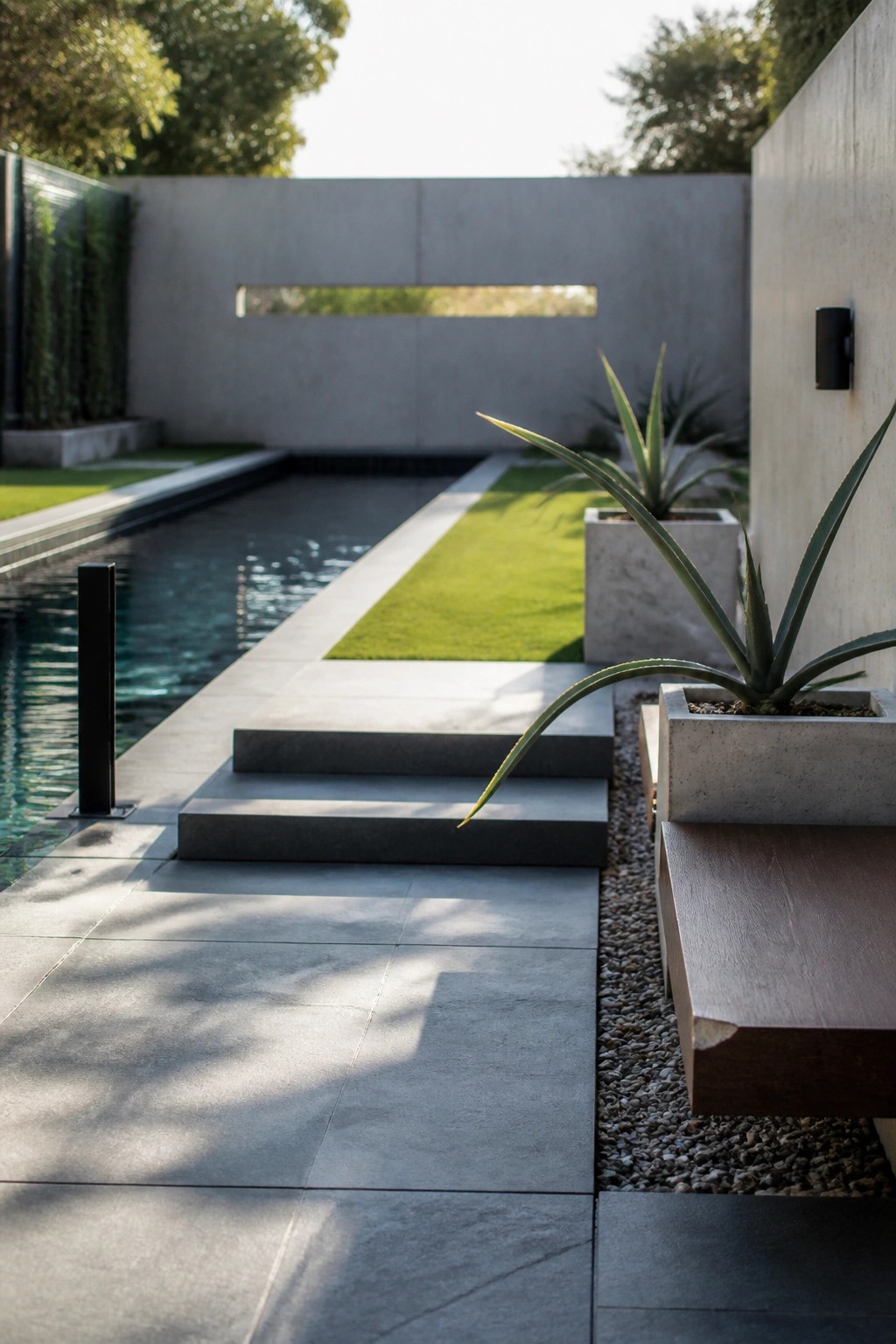 A modern poolside scene shows a long rectangular pool with a concrete deck, steps, a narrow strip of grass, and several concrete planters holding tall agave plants.