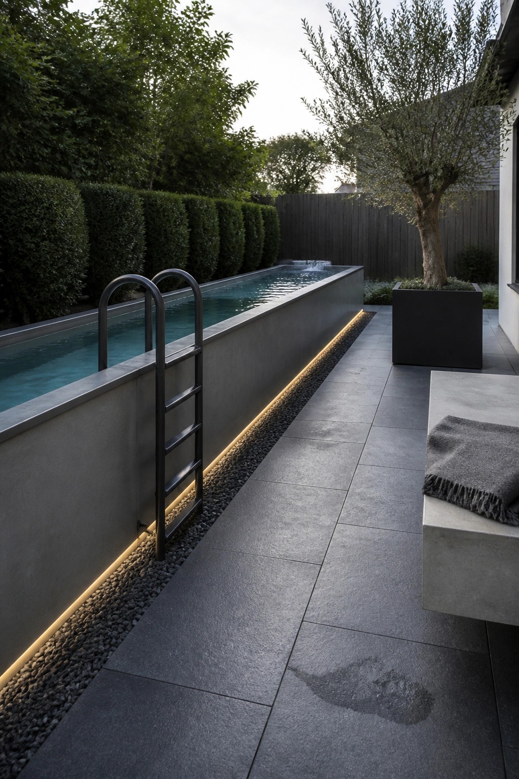 A narrow rectangular pool with a metal ladder is set against a concrete wall with a continuous base light strip, next to a dark paved patio with a concrete bench and large planter.