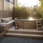 A round wooden above-ground pool on a light wood deck with steps, a built-in bench, and potted plants in a small enclosed yard.