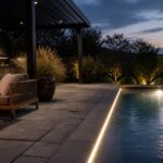 An outdoor pool patio at dusk with a linear light strip along the stone pavers, a fire pit, outdoor sofa, and large potted plants.