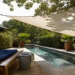 A triangular fabric shade sail is stretched over a swimming pool on a wooden deck, attached to a white post on one side and a tree trunk on the other, with a lounge chair and potted plants in view.