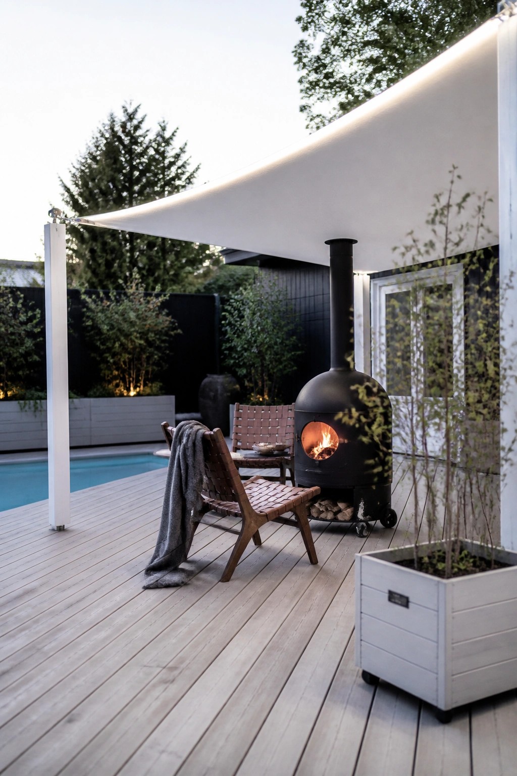 19 Versatile Temporary Pool Shade Ideas for Seasonal Pool Setups A wooden pool deck with a large white shade sail overhead, woven chairs, a black wood stove, and a swimming pool visible on the left.
