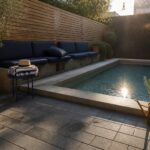 A small rectangular plunge pool with concrete coping set into a gray stone paver patio, next to a built-in bench with blue cushions and several large potted plants.